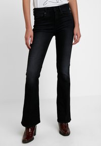 Jeans de mezclilla negros acampanados, con una cintura de media altura y un ligero desgastado. Combinados con botas de tacón marrones.