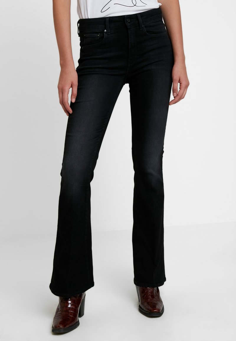 Jeans de mezclilla negros acampanados, con una cintura de media altura y un ligero desgastado. Combinados con botas de tacón marrones.