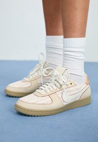 Des pieds portant des chaussettes blanches côtelées et des baskets Nike beige et blanches à motif, avec des lacets noués, se tenant sur une surface bleue.