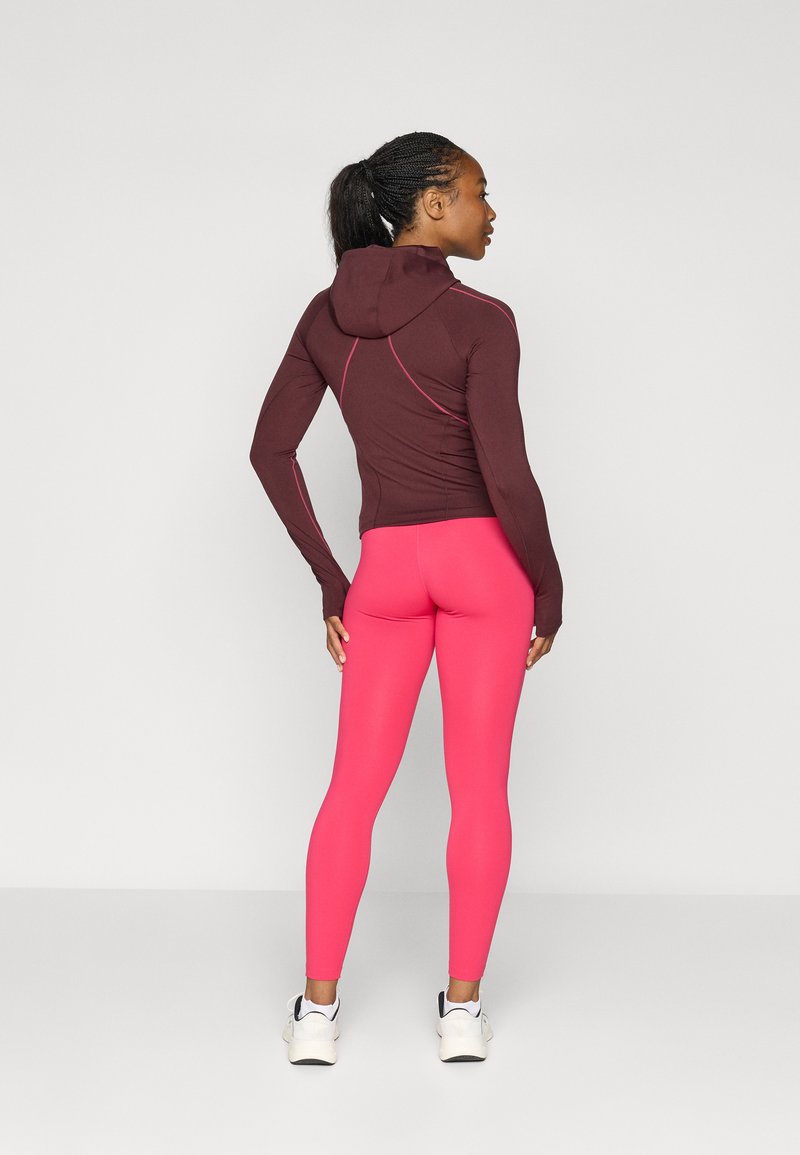 Bordeauxfarbener Kapuzenpullover mit enganliegendem Design und kontrastierendem pinkfarbenem Nähten, kombiniert mit engen pinkfarbenen Leggings. Die Model trägt weiße Sneakers.