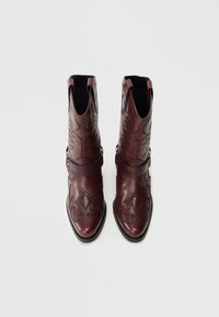 Vīnsarkanas ādas potīšu zābaki ar dekoratīvām šuvēm, smailiem purngaliem un sānu izgriezumiem. Aprīkoti ar vienu siksnu un metāla sprādžu akcentiem.