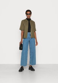 Camisa verde-oliva de manga curta, top preto ajustado, jeans de perna larga azul-claro, sapatos pretos chunky e uma bolsa preta. Os acessórios incluem óculos de sol.