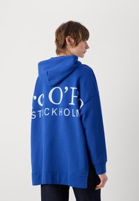 Blauer, übergroßer Hoodie aus Baumwolle mit weißem Text auf der Rückseite, der "STOCKHOLM" liest. Verfügt über eine Kapuze und gerippte Bündchen an den Ärmeln.