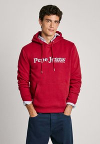 Sudadera roja con un bolsillo frontal y cordones, que presenta un logo blanco de "Pepe Jeans". Se lleva encima de una camisa a rayas azul y blanca.