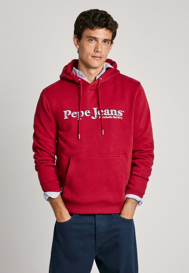 Sudadera roja con un bolsillo frontal y cordones, que presenta un logo blanco de "Pepe Jeans". Se lleva encima de una camisa a rayas azul y blanca.