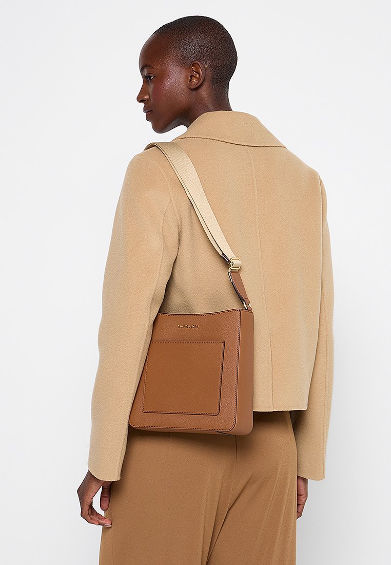 Sac en cuir marron porté à l'épaule avec une poche frontale, des accessoires en métal doré et une bandoulière beige ajustable, porté avec un manteau beige clair.