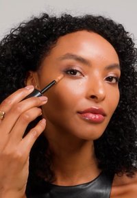 Femme aux cheveux bouclés appliquant un correcteur sous son œil droit à l'aide d'un pinceau applicateur de maquillage, portant un haut noir sans manches.