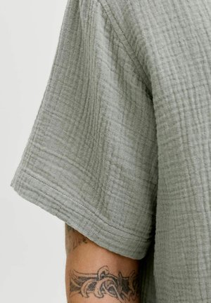 Nærbillede af lysegråt tekstureret kortærmet stof og underarms-tatovering med stammedesign.