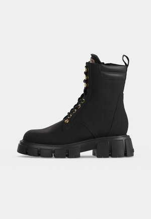 Botte de combat noire à lacets avec semelle épaisse à relief et col de cheville rembourré, vue de côté sur fond gris clair.