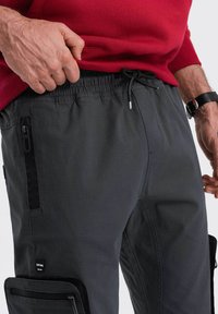 Graue Cargo-Hosen mit strukturiertem Finish, ausgestattet mit einem elastischen Bund, seitlichen Reißverschlusstaschen und einem schwarzen Patch-Logo.