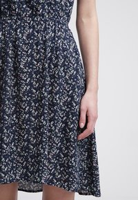 Femme portant une robe bleu marine à petit motif floral, debout avec son bras droit détendu le long du corps.