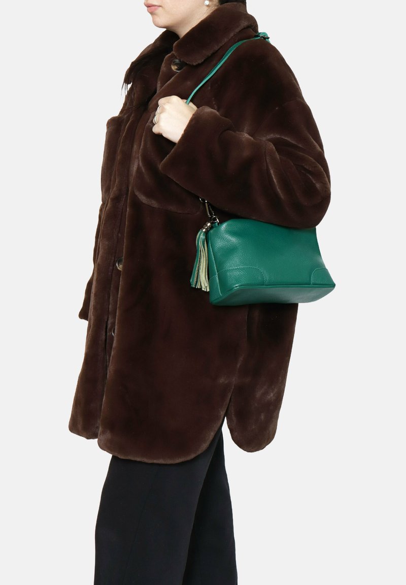 Cappotto in pelliccia sintetica marrone con grandi bottoni, indossato sopra pantaloni scuri. Una borsa in pelle turchese con un pon-pon è tenuta da un lato.
