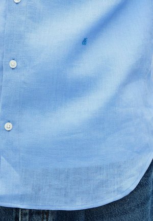 Camisa de lino azul claro con botones blancos, usada sobre jeans de mezclilla azul, mostrando el dobladillo de la camisa y un pequeño logo bordado en el pecho.