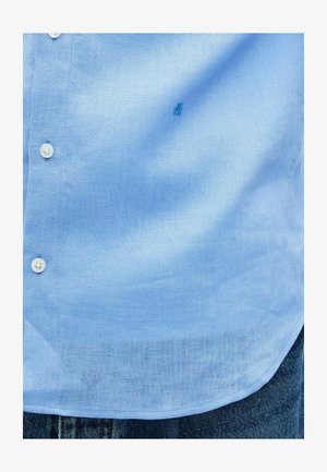 Camicia in lino azzurro con bottoni bianchi indossata sopra jeans di denim blu, con orlo della camicia visibile e piccolo logo ricamato sul petto.