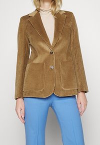 Brun kordfløjl blazer med nålekrave, to frontlommer og en enkelt knaplukning. Sammen med blå skræddersyede bukser.