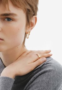 Pendientes de aro geométricos dorados y un anillo dorado con un charm redondo. La modelo tiene el cabello corto y lleva un suéter gris de tela texturada.