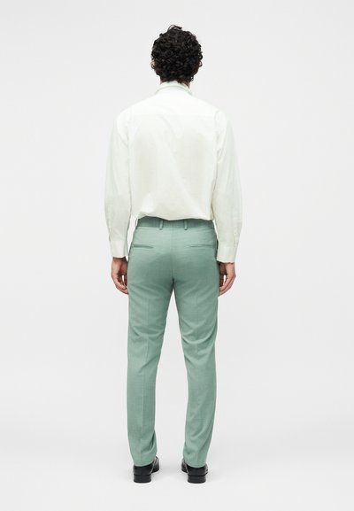 Άνδρας με σγουρά σκούρα μαλλιά στέκεται πλάτη φοράει λευκό πουκάμισο με μακριά μανίκια, ανοιχτό πράσινο παντελόνι και μαύρα παπούτσια σε απλό φόντο.