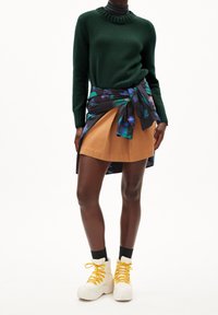 Un pull en maille verte, une jupe évasée couleur beige avec un wrap floral et des baskets blanches à semelles épaisses avec des lacets jaunes. Matériaux texturés et couleurs contrastantes.