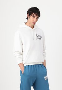 Balta sudegta su gobtuvu, su priekinėmis kišenėmis ir juodu "Calvin Klein" logotipu. Derinama su mėlynomis jogger kelnėmis su CK logotipo pleistru.