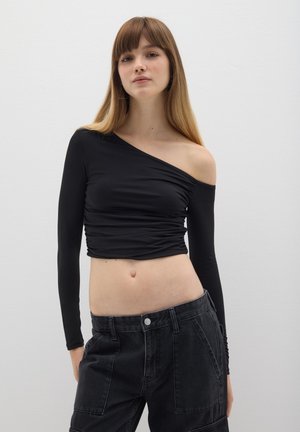 Top nero a maniche lunghe con una spalla scoperta, caratterizzato da un design arricciato e una silhouette aderente. Indossato con pantaloni neri a vita alta.