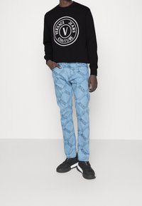 Schwarzer Sweatshirt mit rundem Versace Jeans Couture Logo; hellblaue Denim-Jeans mit wiederholtem dunkelblauem Logomuster; schwarze Turnschuhe.