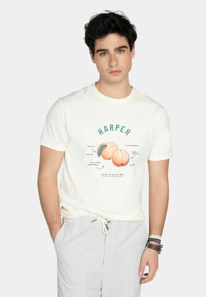 Joven con cabello negro que lleva una camiseta blanca con dos duraznos y la palabra "HARPER" encima, combinado con pantalones claros a rayas.