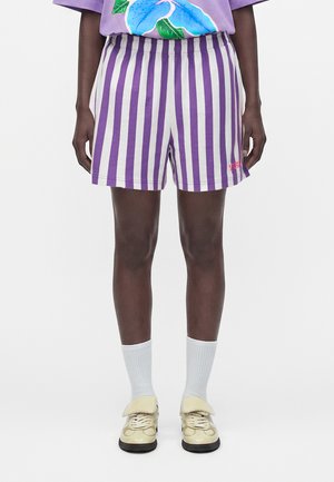 Person iført lilla og hvide vertikalt stripede shorts, hvide sokker, beige sko og en lilla skjorte med et stort blåt blomstermotiv.