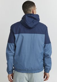 Giacca a vento blu e blu navy con cappuccio, caratterizzata da un design a blocchi di colore, polsini elasticizzati e orlo con cordino. Tessuto morbido e leggero.