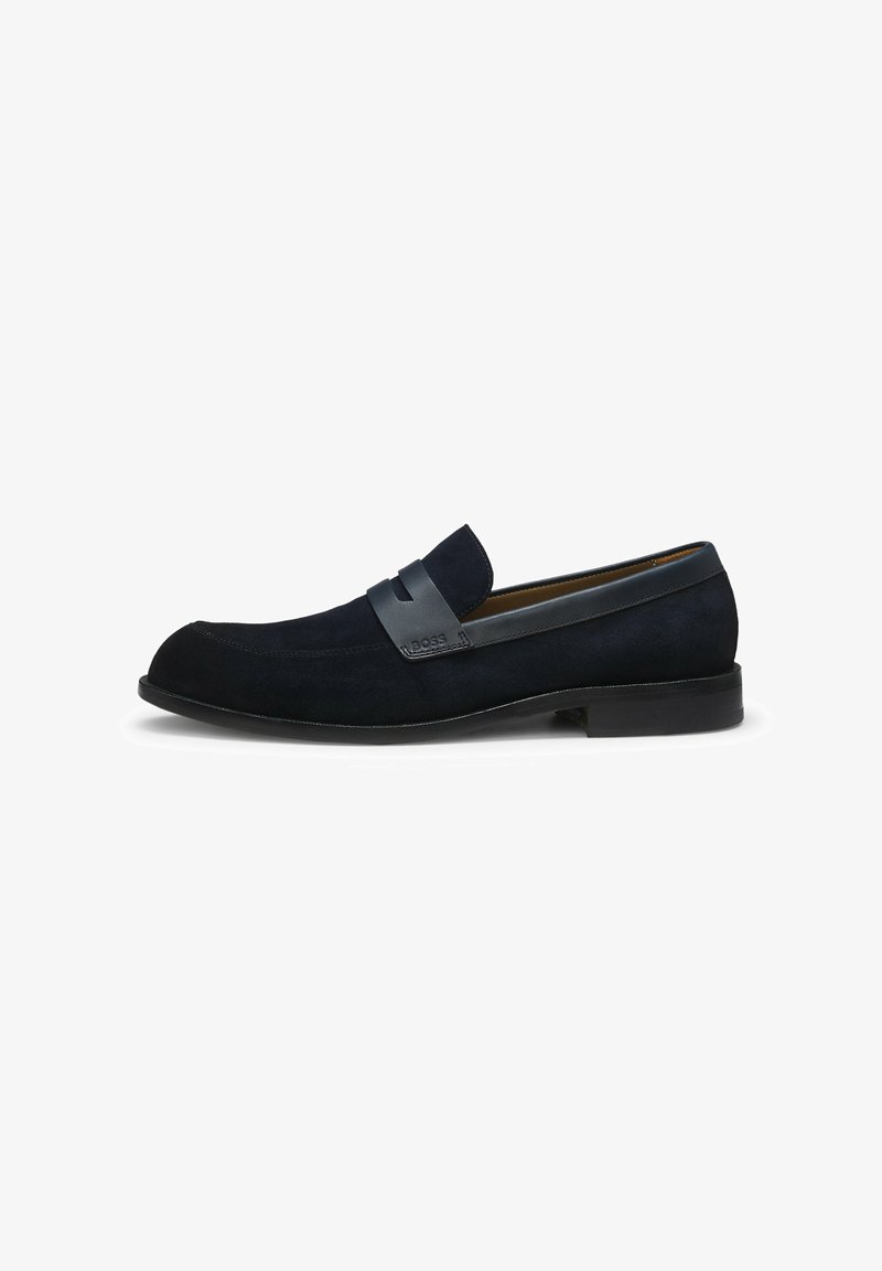 Mocassino slip-on da uomo in suede nero con dettaglio di cinturino in pelle liscia e tacco basso, mostrato di profilo su sfondo bianco.