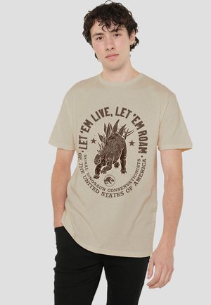 Junger Mann mit lockigem Haar trägt ein beiges T-Shirt mit einem Stegosaurus und einem Text zur Förderung des Dinosaurierschutzes in den Vereinigten Staaten.