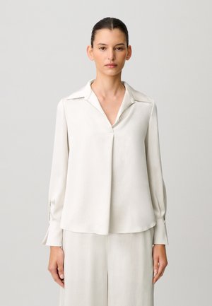 Mujer con cabello oscuro en un moño bajo lleva una blusa blanca suelta de manga larga con puños anchos y pantalones blancos a juego sobre un fondo gris claro.