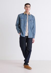 Denim shirt with light brown collar, two chest pockets, and button closure. Paired with dark jeans and tan shoes. Casual fit.