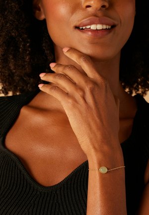 Bracelet en chaîne dorée avec une pierre verte ovale, porté sur une main à la teinte de peau naturelle, à côté d'un haut noir texturé.