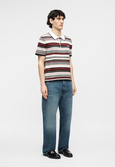 Νεαρός άνδρας όρθιος, φορώντας ριγέ κοντομάνικο πόλο, μπλε τζιν και μαύρα επίσημα παπούτσια, σε απλό ανοιχτό φόντο.