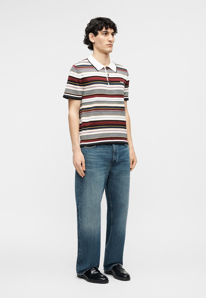 Young man standing, wearing a striped short-sleeve polo shirt, blue jeans, and black dress shoes, on a plain light background.