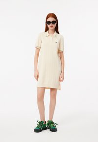 Abito polo beige chiaro con texture a coste verticali, dettagli del colletto in verde e giallo a contrasto, abbinato a sneakers verdi e nere.