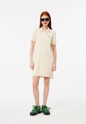 Abito polo beige chiaro con texture a coste verticali, dettagli del colletto in verde e giallo a contrasto, abbinato a sneakers verdi e nere.