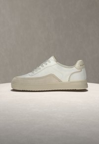 Baskets décontractées blanches et beiges avec laçage à l’avant et semelle plate, affichées sur une surface et un fond neutres.
