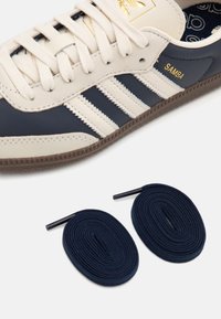 Navy and cream leather sneaker with three white stripes, textured brown outsole, paired with dark blue round laces.