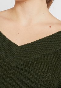 Mörkgrön stickad tröja med v-ringning. Har texturerad ribbstickning och en lös passform. Närbild på halsringning och axelområde.