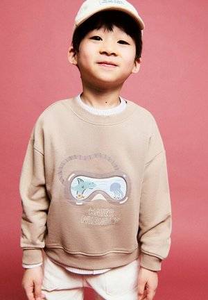 Jeune garçon portant un sweat-shirt beige avec un motif de masque de plongée montrant des créatures marines et le texte « Water Friendly », sur un fond rose.