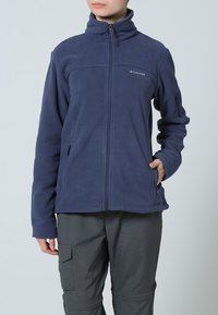 La grâce de l'elegance des styles marins se reflète dans ce blouson en polaire bleu marine, doté d'un col haut, d'une fermeture éclair intégrale et de deux poches latérales. Un logo orne le côté gauche de la poitrine, tandis qu'une finition douce et texturée apporte une touche de confort.