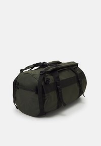 Mørkegrønn duffelveske med svarte stropper, med en rund form, topphåndtak og en sidelomme med glidelås. Holdbart materiale med minimal merking.