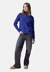 Pull en tricot bleu à col montant, associé à un jean large gris foncé et des chaussures slip-on beiges. Texture lisse et design ajusté.