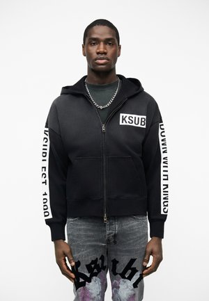 Άνδρας που φορά μαύρο φούτερ με φερμουάρ και την επιγραφή "KSUB" στο στήθος, καθώς και κείμενο στα μανίκια, συνδυασμένο με γκρι τζιν που διαθέτει μαύρα και μοβ γραφικά σχέδια.
