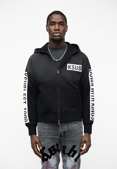 Homme portant un sweat à capuche noir zippé avec "KSUB" sur la poitrine et du texte sur les manches, associé à un jean gris avec des motifs graphiques noirs et violets.