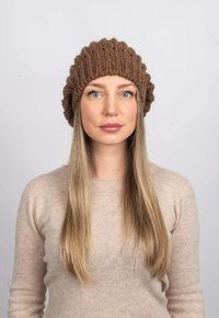 Hnědá pletená čepice s tlustým žebrovaným designem, kterou nosí osoba ve světle béžovém svetru s dlouhými rovnými vlasy. Neutrální pozadí.