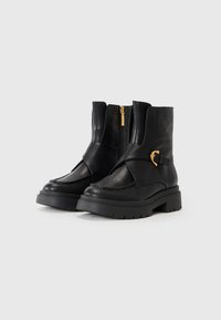 Botine negre din piele cu o platformă pronunțată, având un fermoar lateral și o curea decorativă cu cataramă aurie peste față.