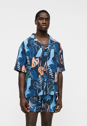Uomo in piedi che indossa una camicia blu navy a maniche corte e pantaloncini con una grande stampa di foglie tropicali blu, arancione e turchese.
