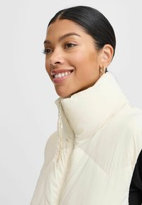 Beige puffer vest met hoge kraag, voorkant met rits en horizontale stiksels. De stof lijkt glad en lichtjes textuur te hebben.